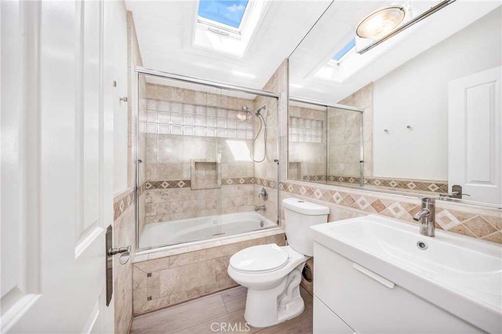 Modern bathroom on Hanover Dr, Costa Mesa, CA, with beige tiles, a bathtub with a glass shower door, a white toilet, and a sink.