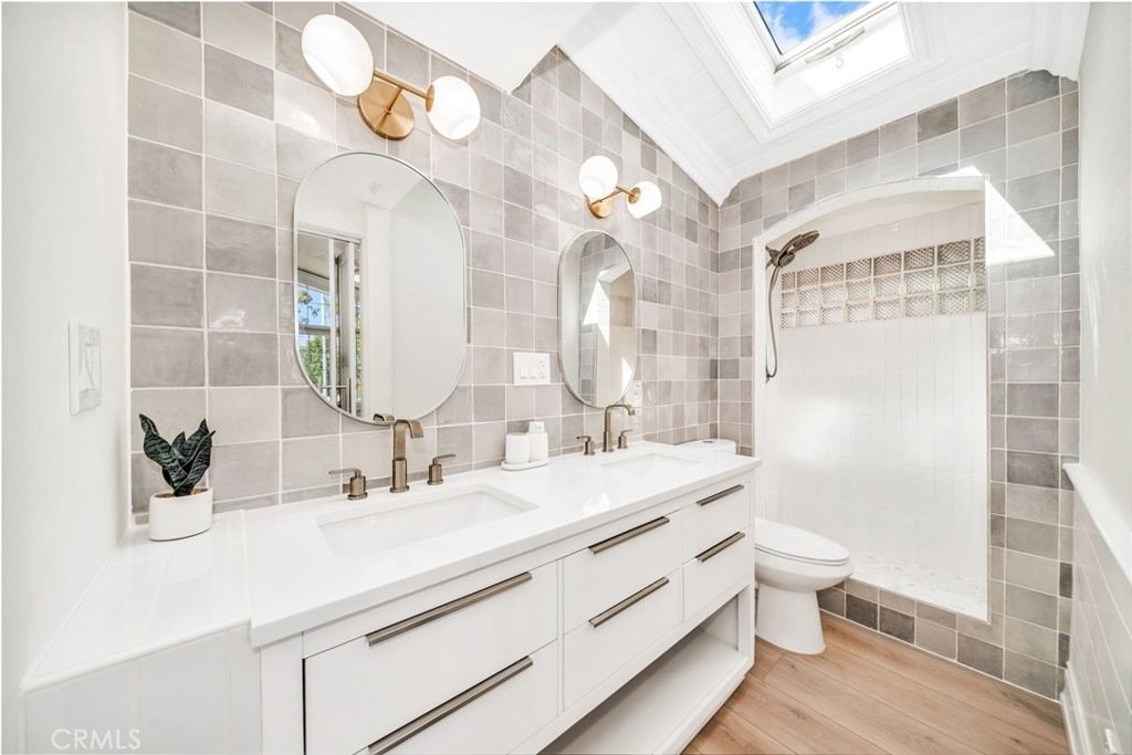 Modern bathroom on Hanover Dr, Costa Mesa, CA, with a skylight, two sinks, oval mirrors, gold faucets, and a gray tiled shower.